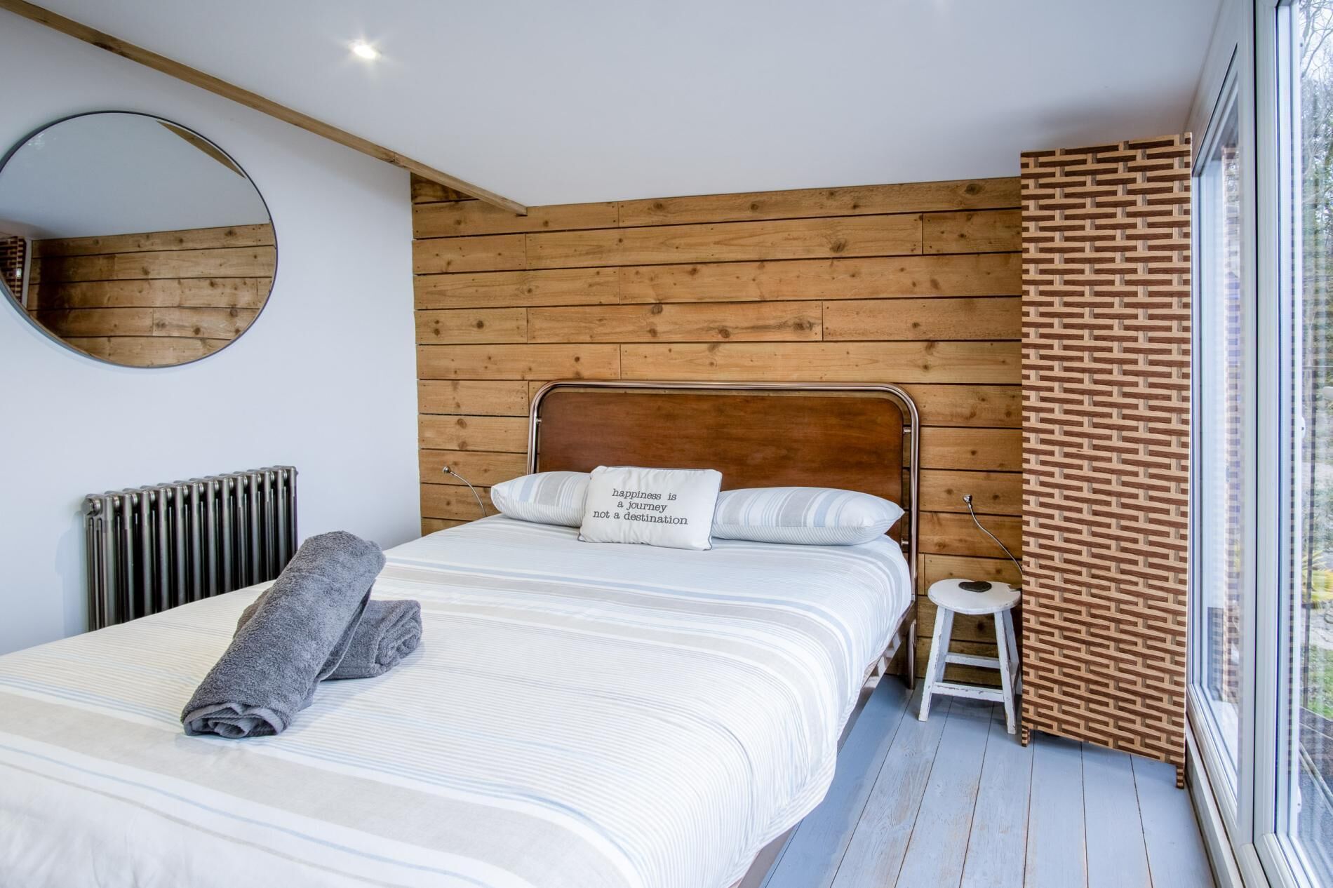 Ein Schlafzimmer mit Holz-Kopfteil, einem runden Spiegel, einem Heizkörper, einem kleinen Hocker und einem Fenster mit Aussicht.