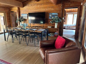 A wooden cabin interior with a dining area, leather couch, and a TV mounted on the wall.