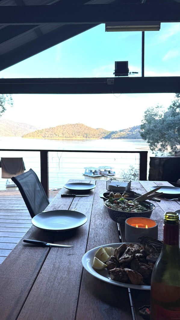 Una mesa de madera con platos, una vela y una botella, con vistas a un lago con barcos y colinas al fondo.