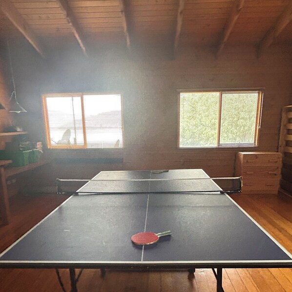 A ping pong table in a room with wooden floors and a sloped ceiling. There are two windows with blinds, and a shelf with various items.