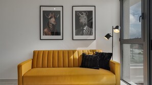 A mustard yellow sofa with a black cushion, two framed animal portraits on the wall, and a floor lamp. - Phaedrus Living: Paphos Noir Residence (Paphos)