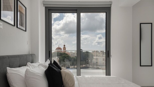 A bedroom with a large window, a bed with white bedding, and a grey headboard. - Phaedrus Living: Paphos Noir Residence (Paphos)