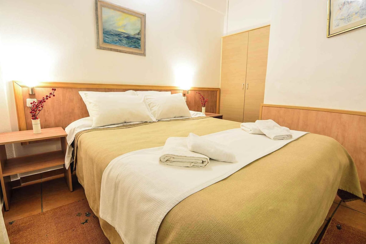 Een keurig opgemaakt bed met witte lakens en een gevouwen handdoek, een nachtkastje met een bloemenvaas, en een houten kledingkast in een hotelkamer.