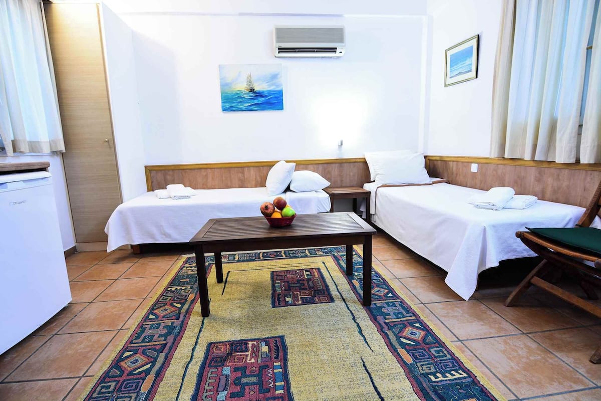 Een hotelkamer met twee bedden, een kleine tafel met fruit, een kamerkleed met patroon en een schilderij van een zeilboot aan de muur.