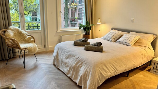 A bedroom with a large bed, a chair, a potted plant, and a view of the outside.