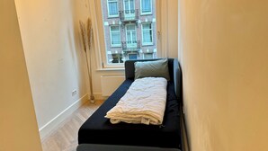 A small, narrow room with a single bed, a window, and a view of a building outside.