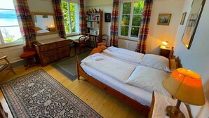 A bedroom with a double bed, a desk, a chair, a bookshelf, and a window with curtains.