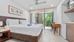 A modern hotel room with a large bed, a ceiling fan, a desk with a chair, and a balcony with a view of greenery.