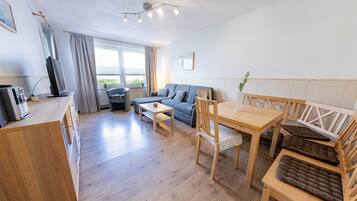 Ein modernes, helles Zimmer mit einem Holzesstisch, einer Couch und einem Fernseher auf einem Schrank.