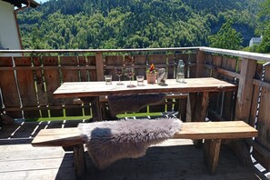 Une terrasse en bois avec une table et un banc, entourée d’une clôture en bois, et offrant une vue sur une colline boisée.