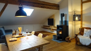 A cozy loft-style living space with exposed wooden beams, a dining table set with plates, glasses, and a decorative deer figurine. A fireplace with a TV above it and a hanging lamp are also visible. - Apartment in Stans near Wolfsklamm River (Stans)