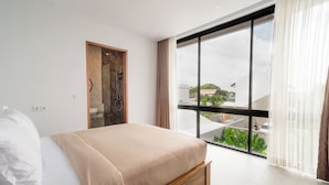 A modern bedroom with a large bed, a wooden door, and a balcony with a view of greenery and buildings. - Sanur Bali Villas by Premier Hospitality Asia (Denpasar)