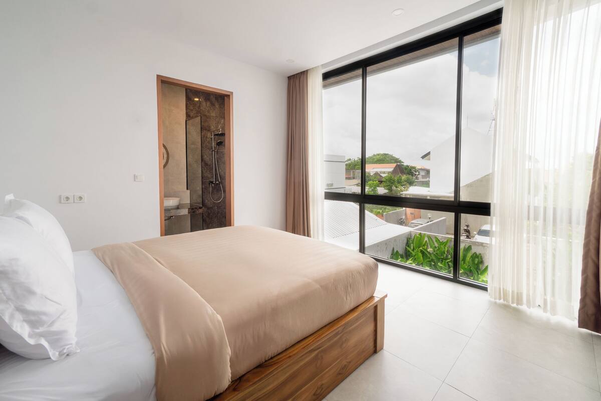 Une chambre moderne avec un grand lit, une porte en bois et un balcon offrant une vue sur la verdure et les bâtiments.