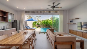 A modern living room with a wooden dining table, sofas, and a TV. There is a pool with a view of greenery outside. - Sanur Bali Villas by Premier Hospitality Asia (Denpasar)