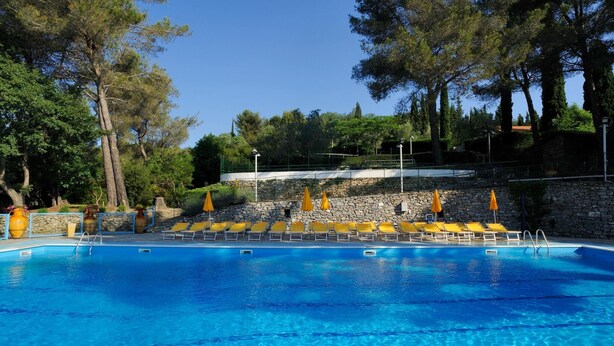 A clear blue swimming pool with yellow lounge chairs and orange umbrellas, surrounded by trees and stone walls.