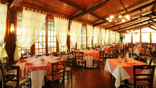Een ruime eetzaal met meerdere tafels, houten balken en grote ramen met gordijnen.