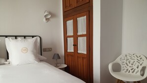 Un lit bien fait, avec un oreiller orné d’un logo, une lampe de chevet, une porte en bois dotée d’une fenêtre grillagée et une chaise blanche au design original.