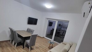 Ein modernes Wohnzimmer mit Holzboden, einem beige farbenen Sofa, einem Esstisch mit Stühlen, einem flachbildfernseher an der Wand und einer Schiebetür zu einer Küchenzeile.