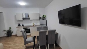 Eine moderne Küche mit weißen Schränken, einem Esstisch mit vier Stühlen, einem flachbildfernseher an der Wand und einer Zimmerpflanze.