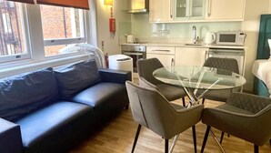 Un espai habitable compacte amb un sofà, una taula de dinar de vidre i cadires, una cuineta amb armaris i electrodomèstics, i una finestra amb persianes.