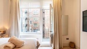 Habitación de hotel con cama, ventana con cortinas y vista a edificios, además de una cesta en el suelo.