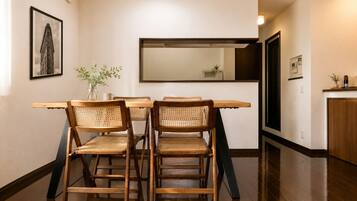 A dining area with a wooden table and four wicker chairs, a vase with a plant, and a framed picture on the wall.