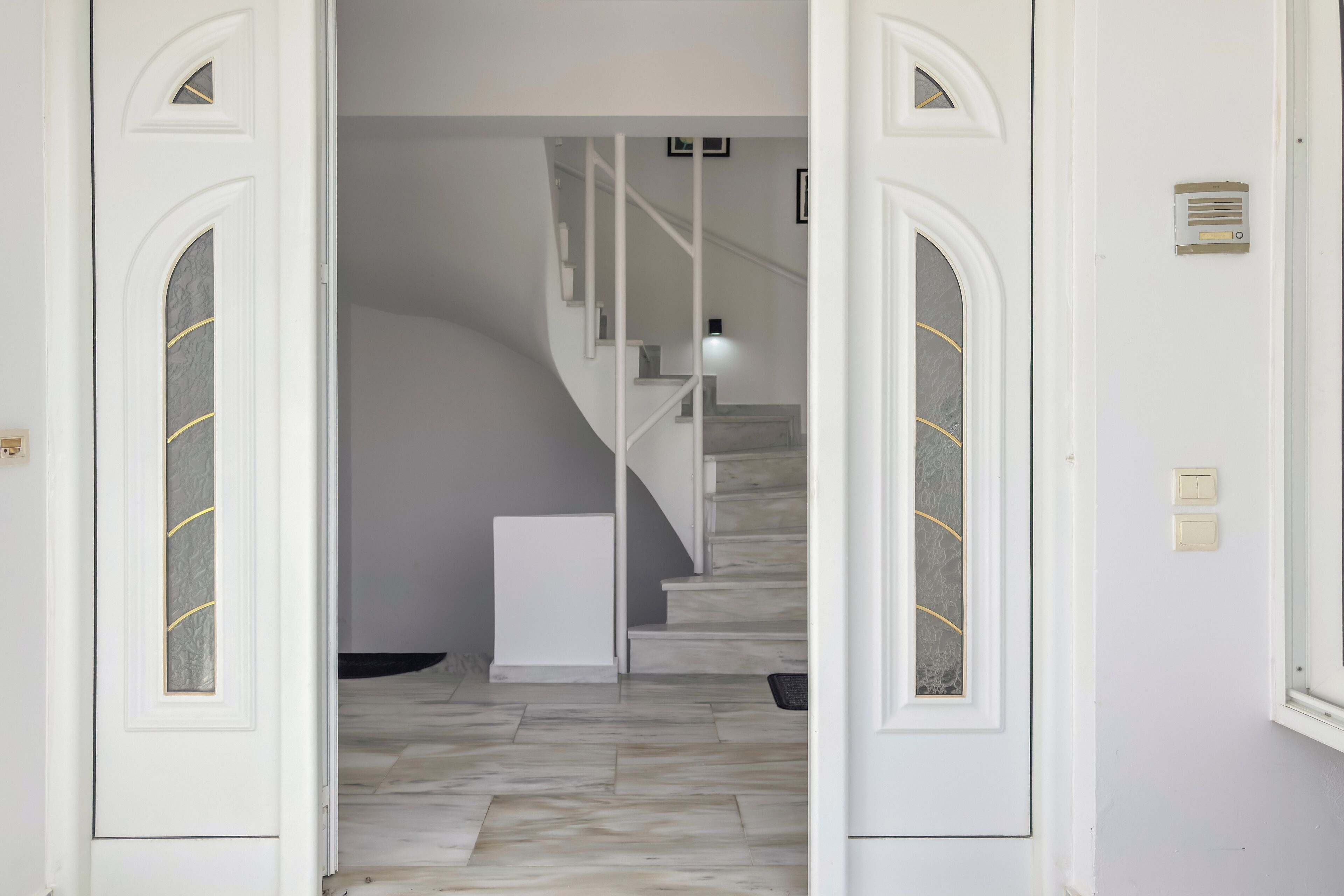 Un pasillo con piso de mármol que conduce a una escalera con barandas blancas y una lámpara colgante.