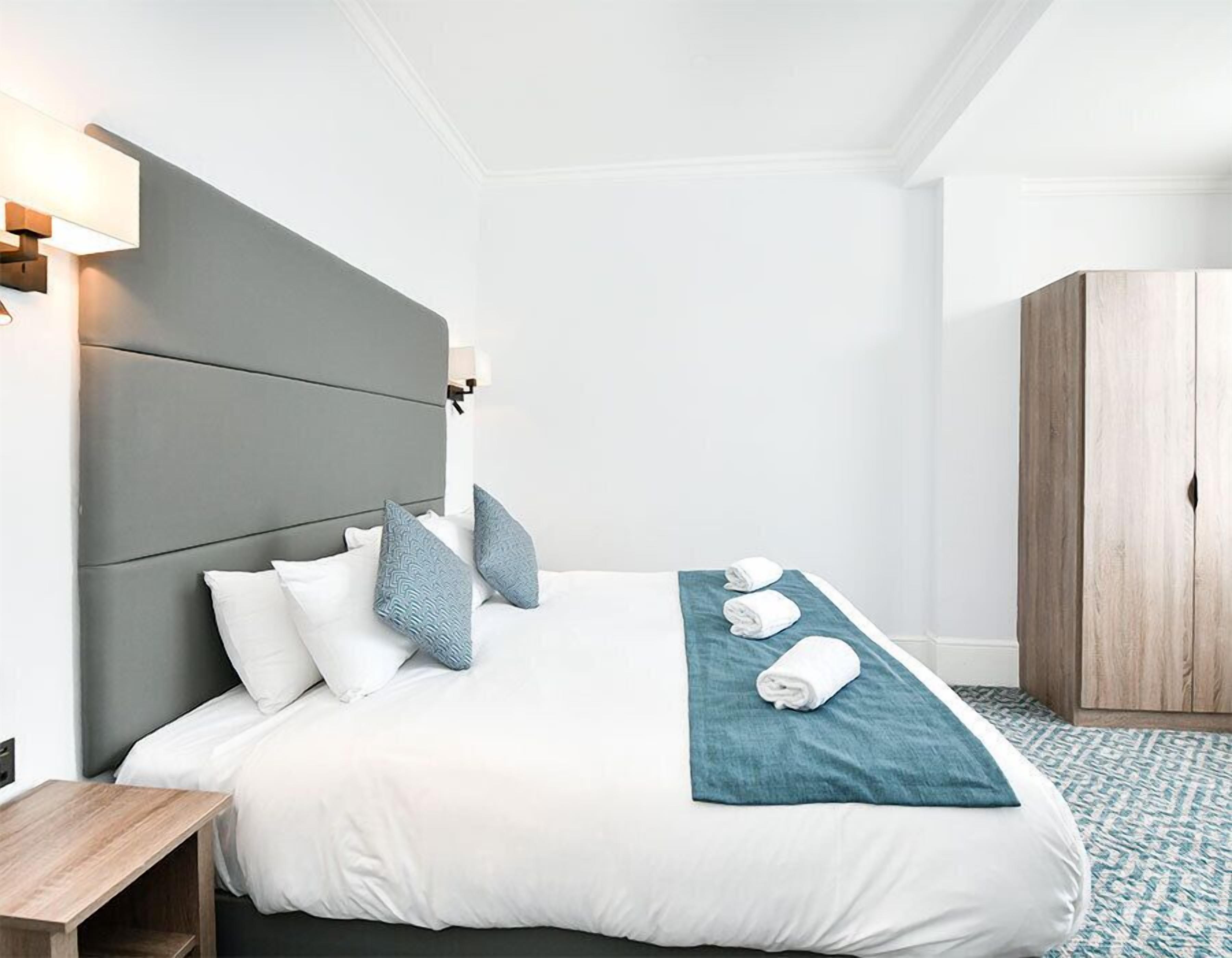 A hotel room with a bed, a nightstand, a wooden wardrobe, and a wall-mounted lamp.