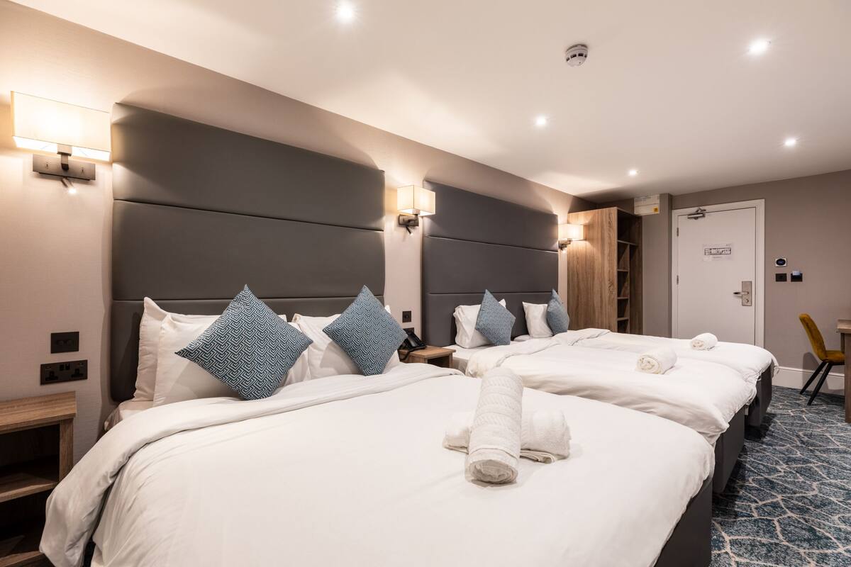 A hotel room with two beds, each with white linens and blue patterned pillows. There are wall-mounted light fixtures and a door with a handle.