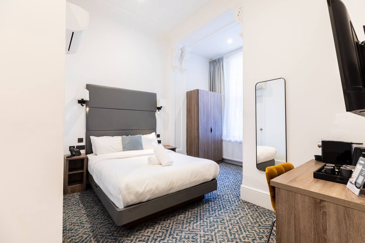 A modern hotel room with a large bed, a desk with a chair, a wardrobe, and a mirror.
