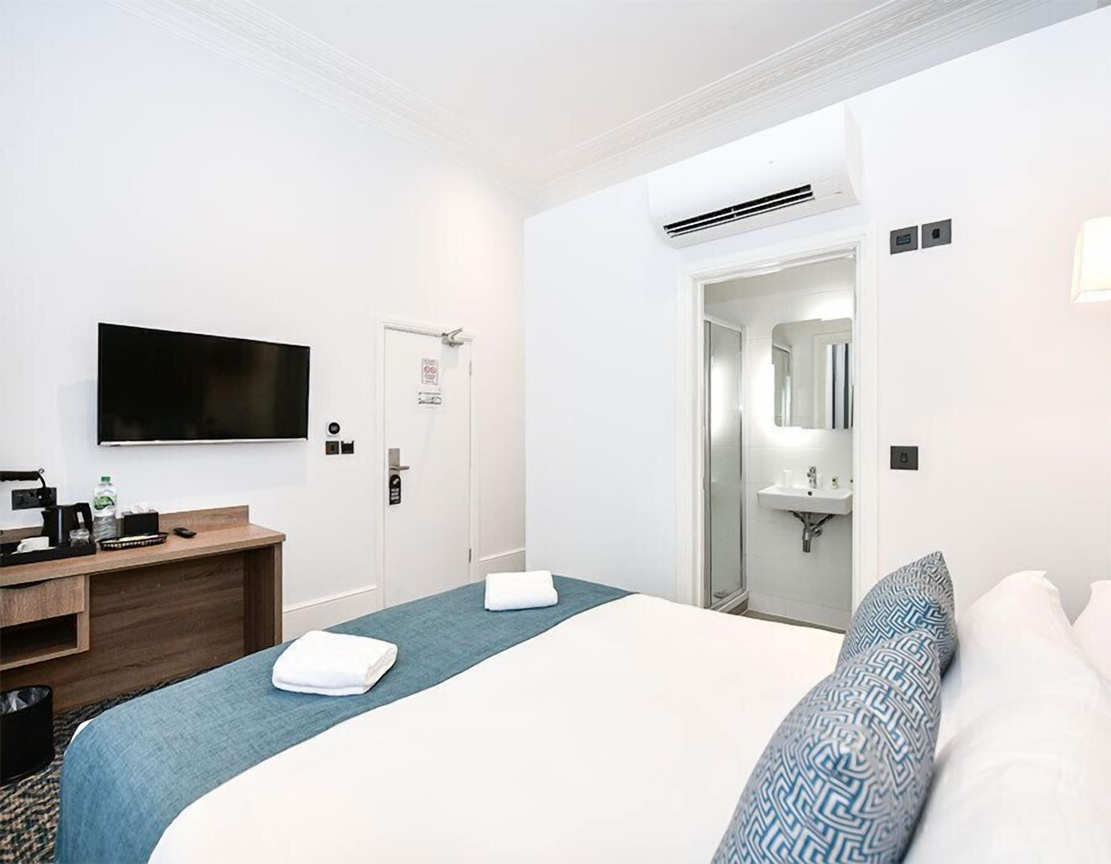 A hotel room with a bed, a desk with a coffee maker, a TV, a shower, and a bathroom mirror.