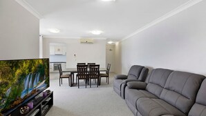 Ett vardagsrum med en grå soffa, en tv på ett tv-bord, ett matbord med stolar och ett kök i bakgrunden.