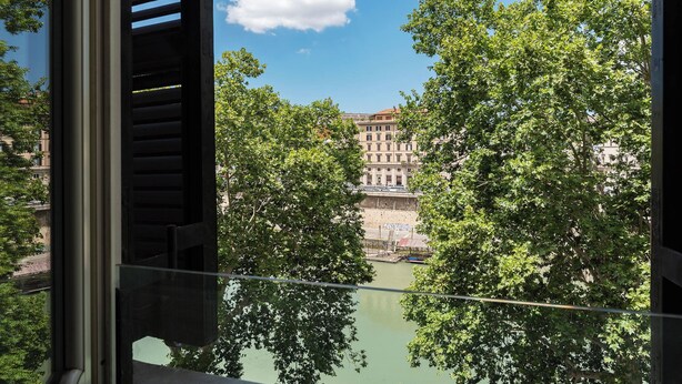 Vista a través de una ventana con baranda de metal, sobre un río y edificios con árboles al frente.