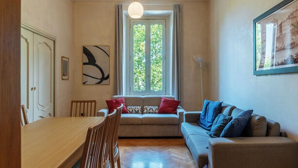 A living room with a wooden dining table, a sofa with blue cushions, and a window with curtains.