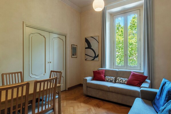 Una habitación con una mesa de comedor y sillas, un sofá con cojines, un ventanal con vista a follaje, y un cuadro abstracto colgado en la pared.
