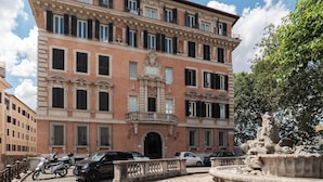 Un edificio de varios pisos con fachada clásica, una entrada central y una fuente al frente.
