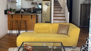 A living room with a yellow sofa, a glass coffee table, and a wooden floor. There is a kitchen area with a microwave, a refrigerator, and a staircase leading to another level. - Charming single-storey house with saltwater pool. Completely renovated in 2022 (Espéraza)