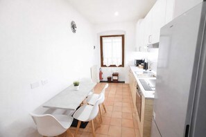 A compact kitchen with white cabinets, a stainless steel refrigerator, a dining table with white chairs, and a window with a white curtain.