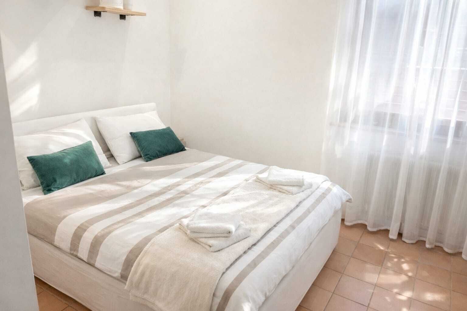 A neatly made bed with striped bedding and two pillows, a wooden shelf above it, and a sheer curtain on the window.