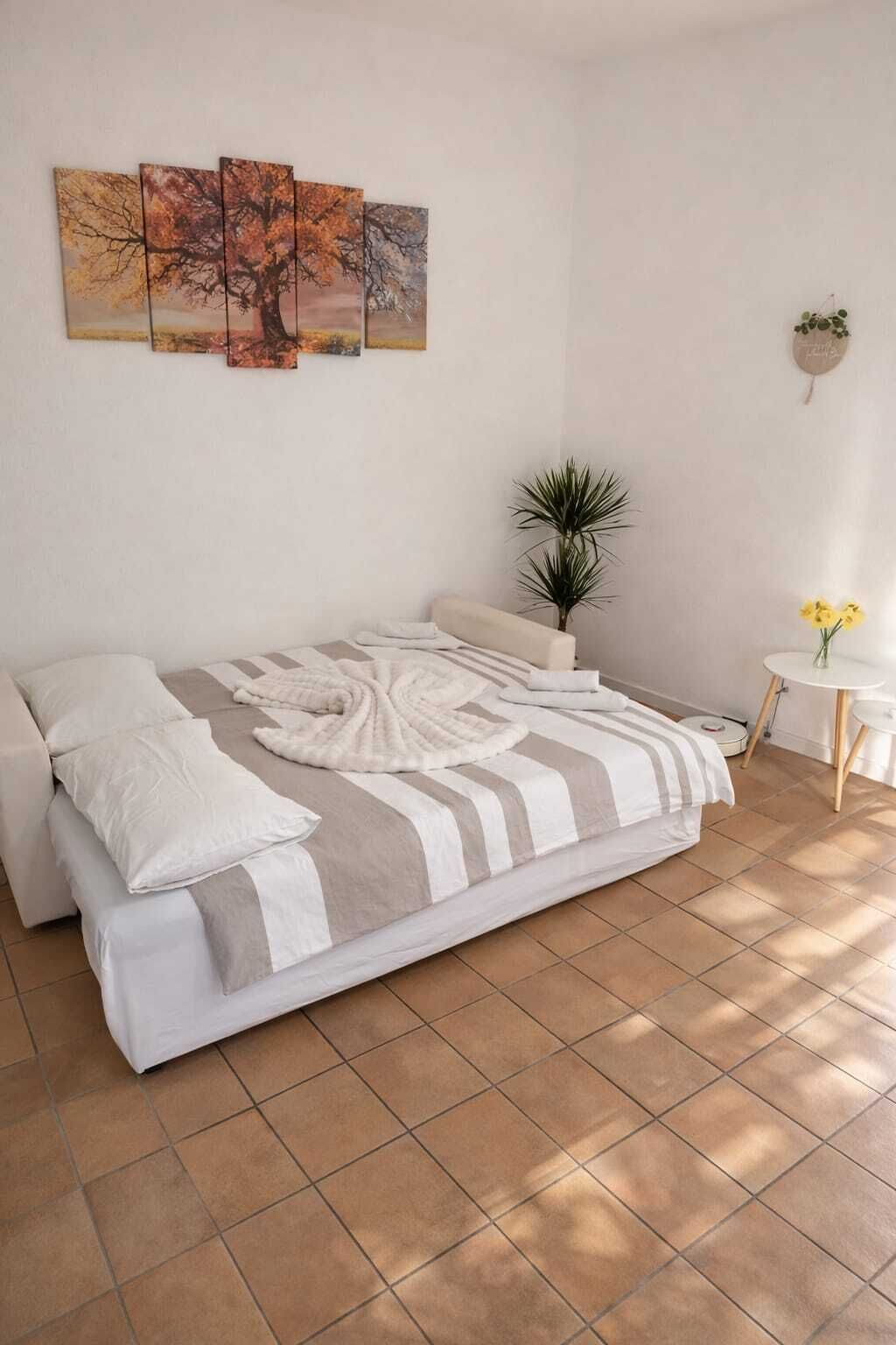 A bedroom with a bed, a potted plant, a small table with flowers, and a wall with three paintings.