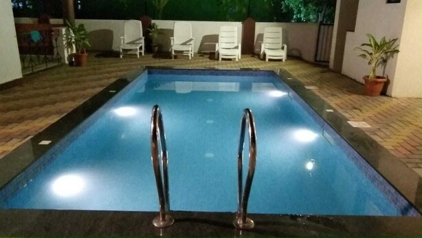 A rectangular swimming pool with two metal ladders at the shallow end, surrounded by white lounge chairs and potted plants. - Rihan Goa Holiday Homes (Calangute)