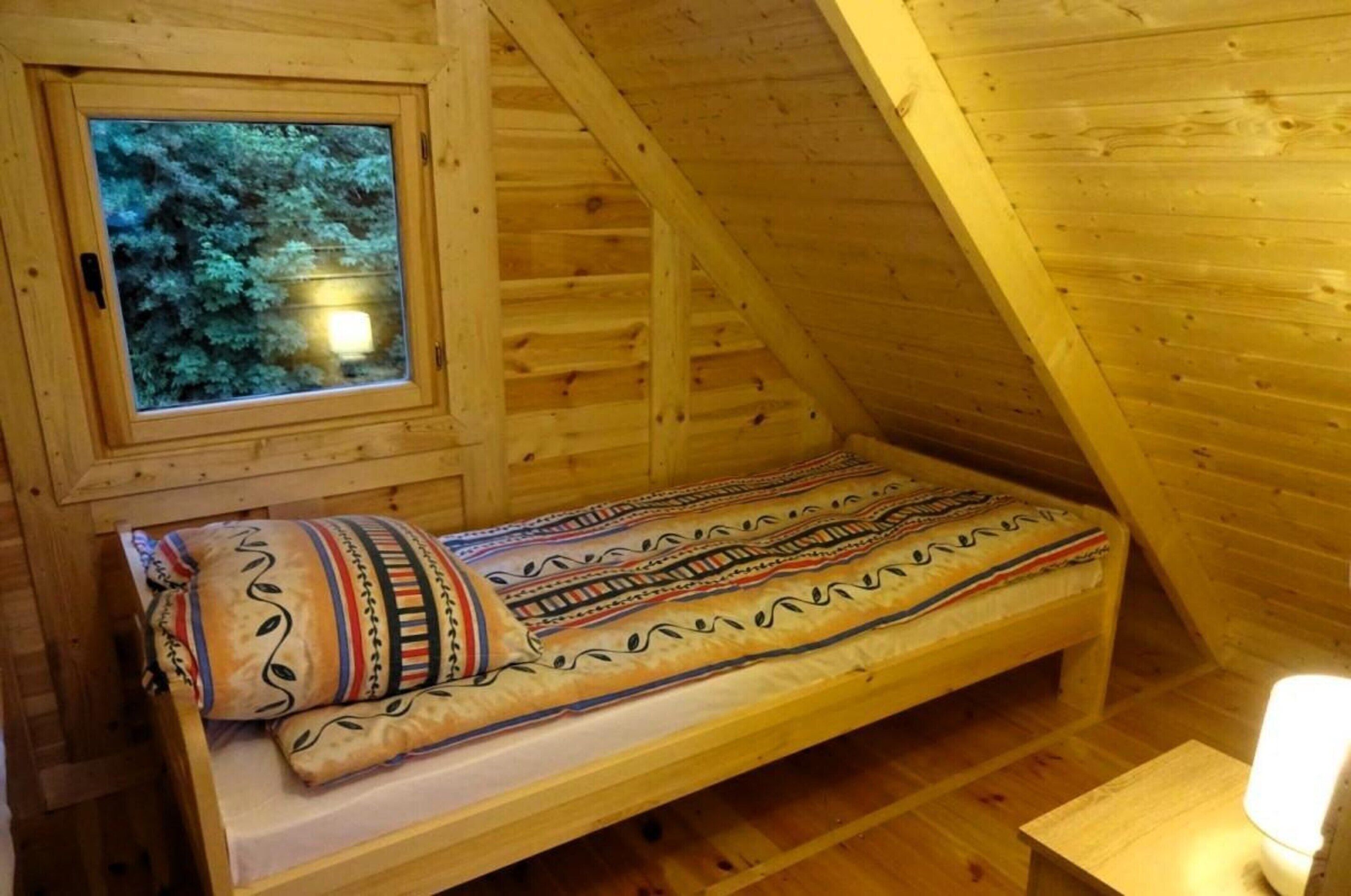 A wooden cabin with a single bed, a window with a view of trees, and a small table with a light.