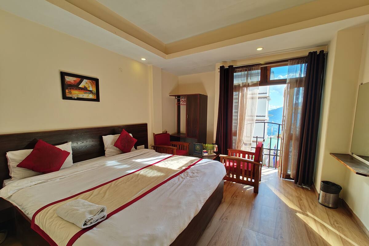 A hotel room with a large bed, a wooden headboard, a red chair, a small table, and a view of the outdoors through a window with curtains.