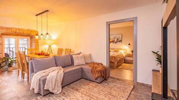 A modern living room with a sectional sofa, a dining area with a brick fireplace, and a bedroom with a bed and bedside table.