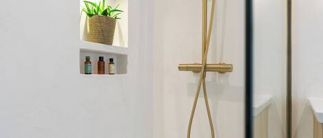 A shower with a gold hose and a wall-mounted control. There is a shelf with a woven basket and three bottles below a window.