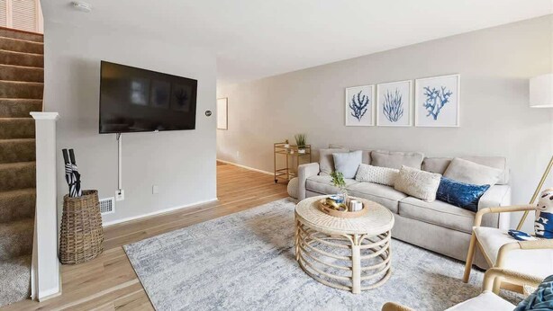 Een moderne woonkamer met een flatscreen televisie aan de muur, een grijze bank met decoratieve kussens, een ronde rieten salontafel, en een trap met houten treden.