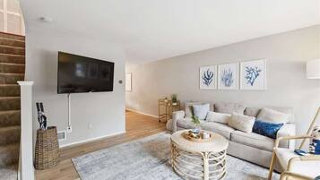 A modern living room with a flat-screen TV mounted on the wall, a grey sofa with decorative pillows, a round wicker coffee table, and a staircase with wooden steps.