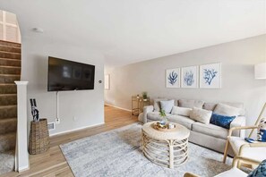 A modern living room with a flat-screen TV mounted on the wall, a grey sofa with decorative pillows, a round wicker coffee table, and a staircase with wooden steps. - Annapolis Blue and Gold Haven (Annapolis)