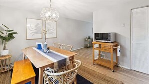 Een eetgedeelte met een houten tafel, rieten stoelen, een hanglamp en een magnetron op een karretje.