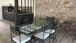 A dining area with a glass table and metal chairs, a kitchen in the background, and a stone wall.